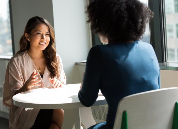 Two women are sitting in a semi-bright room in white chairs across each other at a white desk. The woman facing the screen has long brown hair and is explaining something to the woman across from her who has extremely curly hair and is wearing a blue shirt.
