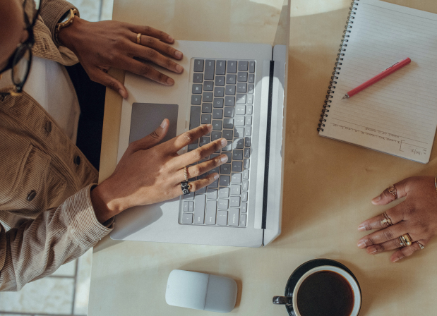 A view from above, two people are sitting at a desk. On the desk is a laptop, a mouse, a notebook with a pencil, and some coffee in a mug. The laptop is being used by one of the people in the photo who is wearing a light spring jacket and glasses.