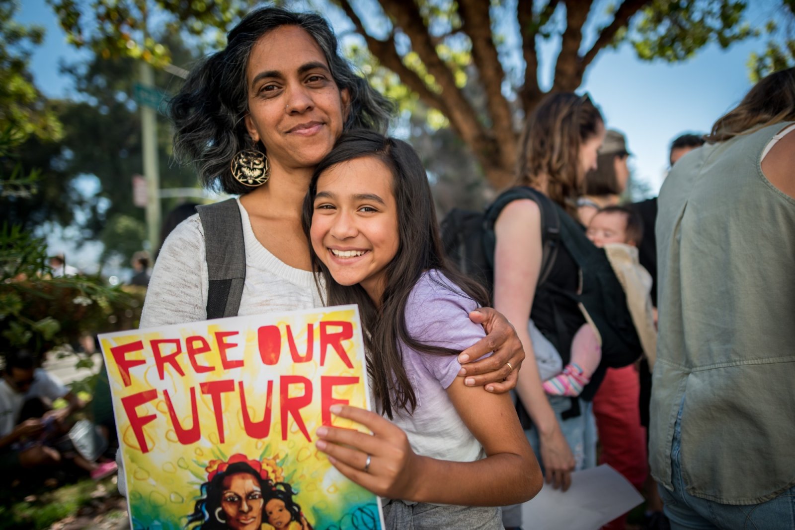 An older woman is holding a younger girl while both of them look directly at the camera and smiling. They are participating in a protest and are holding a sign that says free our future. They are outside in the daytime.
