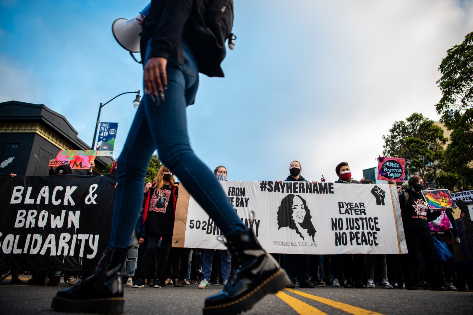 The scene is of a protest in the middle of the street with the view angled from the street level. A person speaking into a microphone is walking across the photo and behind them is a sea of people holding a white banner that says No justice, no peace and #sayhername. Another black sign on the left says in large white lettering Black and Brown solidarity