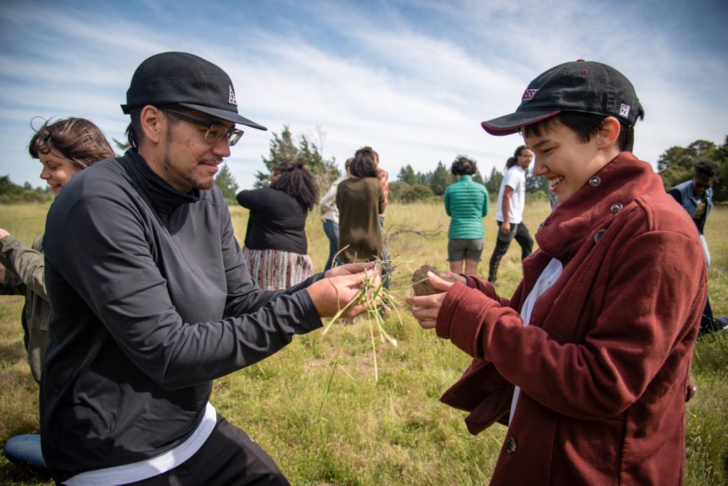 A man in a back jacket and a black hat is handing a pile of grass or hay while smiling to the person in front of him wearing a red jacket and a black hat. They are both standing in a field of grass and are participating in an activity.