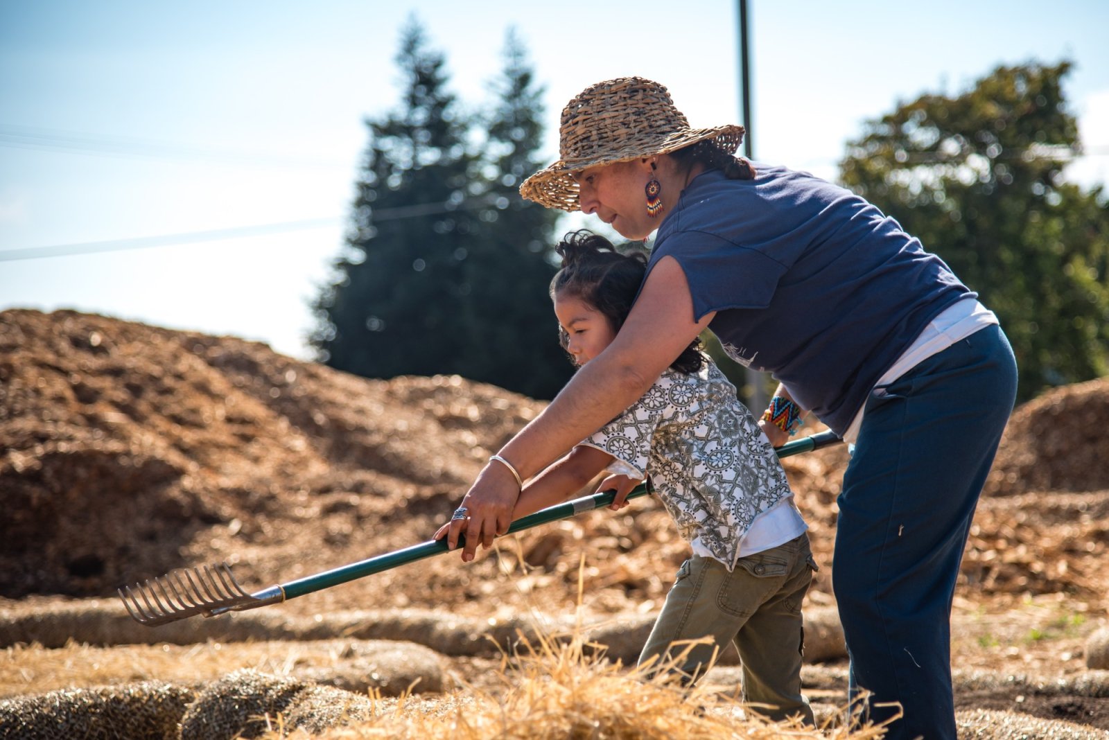 A woman wearing a sun hat and a blue shirt stands in a field with her daughter. She is showing her young daughter a farming technique by helping her hold a long piece farming equipment