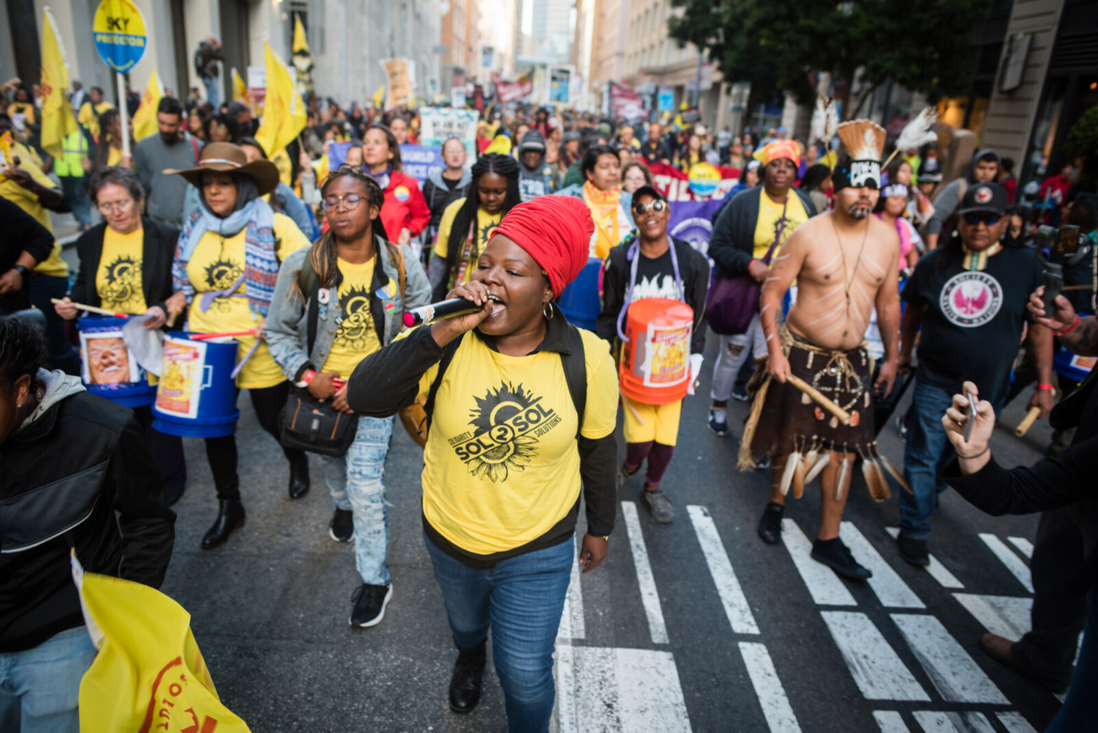 A crowd of people are walking in the middle of the street as part of a protest. Many are wearing a bright yellow shirt as part of the same organization. They are crossing a crosswalk and one person leading the way in the photo is wearing a red hair wrap speaking into a microphone.